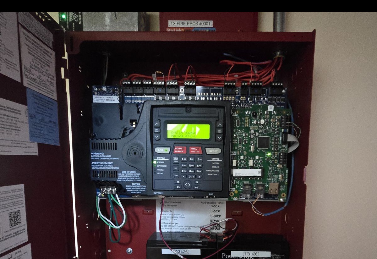 Open addressable fire alarm control panel with field wiring landed on its terminals, a TX Fire Pros-tagged cellular communicator above, and dated standby batteries