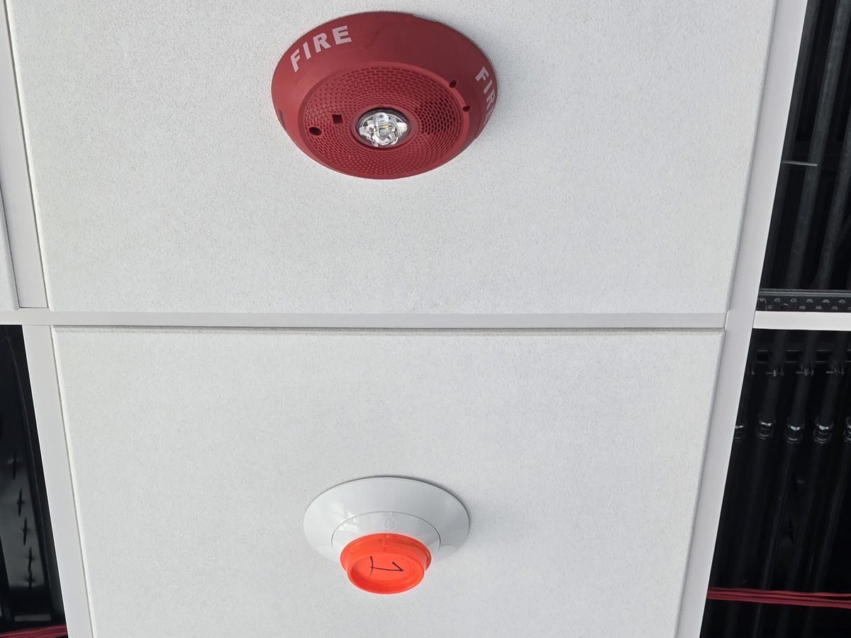 Ceiling-mounted horn/strobe and smoke detector installed in adjacent acoustic ceiling tiles, the detector's protective dust cover still fitted